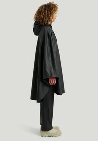 Svart läderponcho med huva, översized design, asymmetrisk fåll, bärs över åtsittande svarta byxor och kraftiga beiga skor.