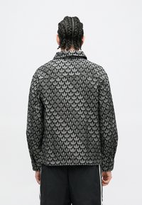 Asmuo su pintinėmis šukuosenomis, dėvintis ilgomis rankovėmis juodai pilką raštuotą švarką ir juodas kelnes su baltomis šoninėmis juostomis, žiūrintis į šoną.