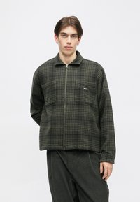 Fleece jakna temno zelene barve s karirastim vzorcem, z zadrgo spredaj in dvema prsoma žepoma. Mehka tekstura z ovratnikom.