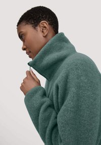 Fleece zip-up jacket in teal, featuring a high collar and relaxed fit. Material appears soft with a subtle texture and embroidered logo on the sleeve.