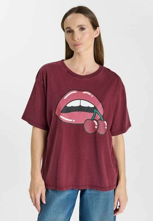 Frau trägt ein locker sitzendes dunkelrotes T-Shirt mit einer Grafik von offenen Lippen, die zwei Kirschen halten, kombiniert mit blauen Jeans, vor einem schlichten Hintergrund.