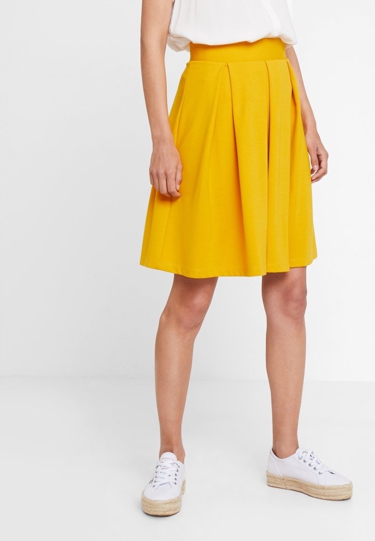 Femme portant une jupe plissée jaune vif longueur genou, un haut blanc sans manches et des baskets espadrilles blanches avec semelles en corde.