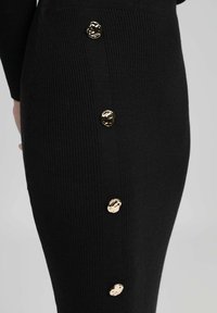 Robe noire côtelée avec cinq boutons décoratifs dorés sur le côté. Le tissu a une texture lisse et une silhouette ajustée.