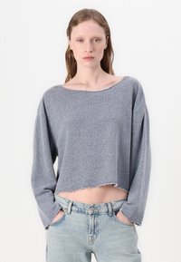Sweater cinzento cortado em tecido de malha macia, apresentando um decote largo, ombros caídos e bainha desfiada. Combinado com jeans azul claro.
