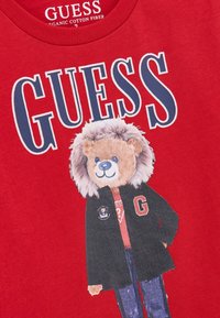 Crvena pamučna majica s grafikom medvjeda s detaljima krzna koji nosi crnu jaknu i plave traperice, s natpisom "GUESS" iznad.