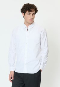 Nuori mies, jolla on kiharat tummat hiukset, valkoinen nappipaita ja mustat housut, seisoo yksinkertaisen valkoisen taustan edessä.