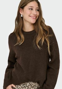 Junge Frau, die lächelt, einen dunkelbraunen Pullover, silberne Ohrringe und Leoprint-Hosen trägt, seitlich blickend vor einem schlichten Hintergrund.