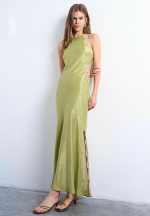 Kvinde i en ærmeløs olivengrøn satin kjole med beige blonde detaljer, stående barfodet mod en hvid væg og grå gulv.