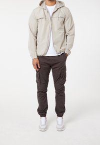 Ljusbeige zip-up jacka med huva, elastiska muddöppningar och två framfickor; matchad med mörka cargobyxor och vita sportskor.