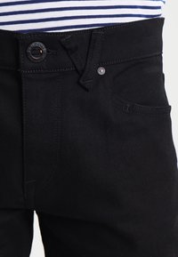 Primer plano de la cintura de unos jeans negros con detalles de botón y remaches, llevados con una camisa a rayas azul y blanca metida por dentro.