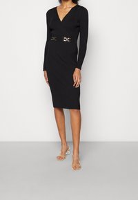 Schwarzes, geripptes Wickelkleid mit langen Ärmeln, V-Ausschnitt und taillierter Passform, akzentuiert durch einen goldfarbenen Gurt mit Schnalle. Glatte Textur und knielang.