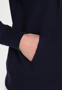 Main insérée dans la poche d'un sweat à capuche à manches longues bleu marine foncé avec poignet côtelé, visible de la taille au-dessus.