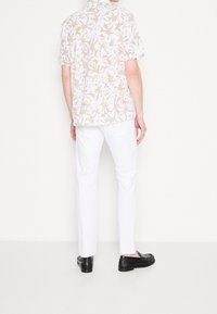 Camicetta bianca a maniche corte con motivo floreale beige, abbinata a pantaloni bianchi e mocassini neri, che mostra un design dal taglio rilassato.