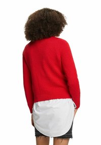 Personne aux cheveux bouclés portant un pull rouge texturé superposé à une chemise blanche et une jupe noire effilochée, vue de dos.