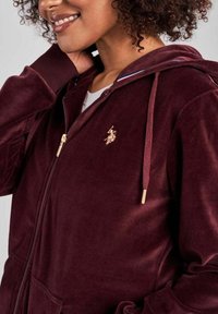 Personne souriante portant un sweat à capuche zippé bordeaux avec des cordons de serrage et un petit logo doré brodé sur la poitrine.