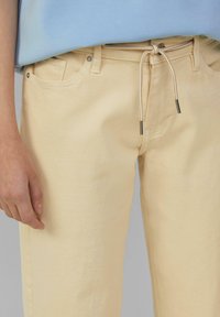 Pantaloni in denim giallo con vita regolabile e accessori in argento. Presentano tasche frontali e una superficie liscia e opaca.