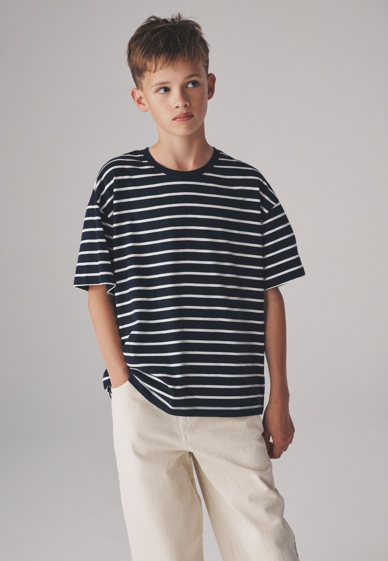 Garçon portant une chemise à manches courtes rayée noir et blanc et un pantalon clair, debout avec une main dans la poche, regardant sur le côté.