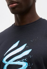 Κοντινό πλάνο ενός ατόμου που φοράει σκουρόχρωμο πουκάμισο με ένα μπλε, αφηρημένο σχέδιο που μοιάζει με νερό στο στήθος.