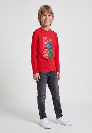 Leende pojke med blont hår som bär en röd långärmad tröja med färgglada Ninjago-figurer, mörka slitna jeans och vita sneakers, stående.