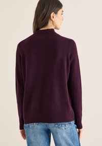 Bordeauxroter Strickpullover mit hohem Kragen, gerippten Bündchen und Saum. Weiche Textur, leicht lockere Passform, kombiniert mit hellblauen Jeans.