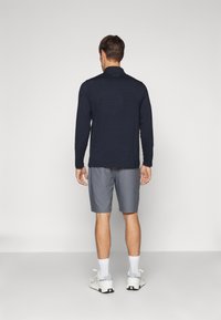 Marineblå langermet skjorte med krage, kombinert med lysegrå shorts. Modellen har på seg hvite sneakers med svart såle og hvite sokker.