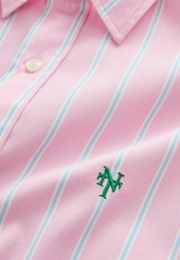 Chemise rose boutonnée avec des rayures verticales bleues et blanches et un logo vert brodé près de la poitrine, montrant le col et le bouton supérieur.