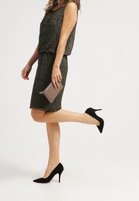 Robe sans manches vert foncé avec un motif géométrique texturé, associée à des escarpins noirs à bouts pointus et une pochette brun clair.