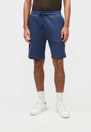 Mężczyzna w granatowych szortach na sznurki oraz białych trampkach z białymi skarpetkami, stojący na jasnym, jednolitym tle.