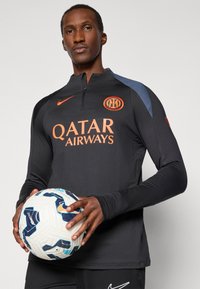 Nike dugulja za trening dugih rukava u crnoj boji s narančastim logotipom i tekstom "Qatar Airways". Model drži nogometnu loptu s uzorkom i plavim akcentima.