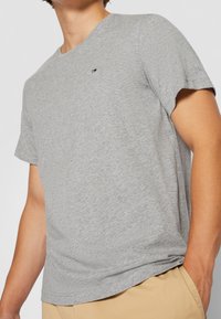 Uomo che indossa una maglietta a maniche corte grigia chiara con un piccolo logo rosso, bianco e nero sul petto, abbinata a pantaloni beige.