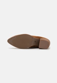Suela de zapato de tacón de ante marrón con un lado de goma texturizada, que presenta patrones estriados y pequeños remaches circulares para mayor tracción.