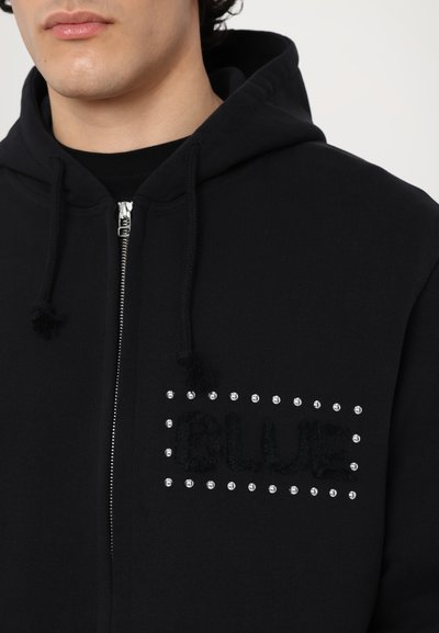 Veste à capuche zippée noire avec une texture douce. Présente un graphique "BLUE" en noir texturé et des strass clairs sur la zone de la poche avant.