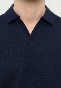 Κοντινό πλάνο ενός ατόμου που φοράει μπλε σκούρο πλεκτό πουκάμισο με υφή, με ανοιχτό γιακά και κοντό μυτερό γιακά.