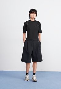 Camisa atlética preta de mangas curtas, combinada com calções largos pretos. O conjunto apresenta costuras cinzas sutis e ténis metálicos.