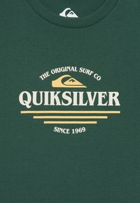 Žalias medvilninis marškinėliai su „Quiksilver“ logotipu balta ir geltona spalvomis, su kreiviniais ir tiesiais tekstais. Klasikinio apvalaus kaklo dizainas.