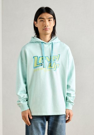 Mint green hoodie with a front graphic reading "Level Up" in blue and yellow. Features a drawstring hood and long sleeves. Worn with blue jeans.