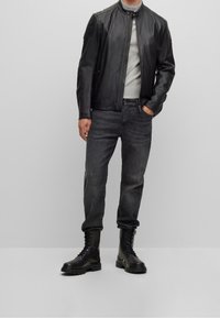 Svart läderjacka med hög krage, dragkedja och axelaccenter; bärs över en ljusgrå tröja och mörka jeans, tillsammans med svarta stövlar.