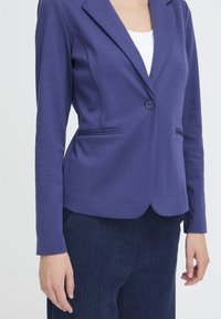 Ženska oblečena v enospojni vijolični blazer, beli majici in temno modri kord pantalooni, stoji s sproščenimi rokami ob straneh.