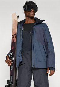 Tengerészkék síkabát texturált panelekből, fekete felsőre rétegezve, sötét sínadrággal párosítva. Kezében sílécet tart feketés kötéssel és sígóllal.