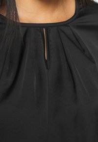 Svart blus med plisserad nacke och en liten nyckelhålsöppning. Tillverkad av ett mjukt tyg med en subtil lyster och utan synlig hårdvara.