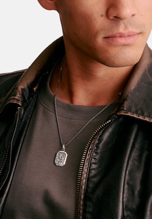 Mann trägt eine dunkle Lederjacke, ein graues Hemd und eine silberne religiöse Anhängerkette mit einer eingravierten Heiligenfigur.