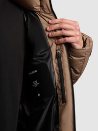 Doorgestikte jas met een bruine buitenkant en een zwarte voering. Voorzien van een rits, een zijzak en een merkembleem aan de binnenkant.