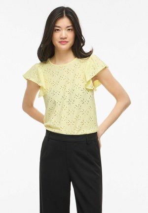 Jeune femme aux cheveux foncés portant un chemisier jaune pâle en œillets avec des manches volantées et un pantalon noir taille haute, les mains derrière le dos.