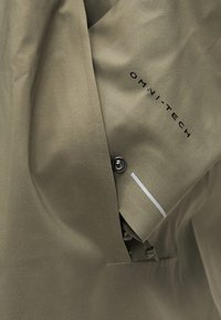 Κοντινό πλάνο υφάσματος χακί με το κείμενο "OMNI-TECH", ένα μεταλλικό κουμπί σναπ και μια λευκή ανακλαστική λωρίδα σε μανσέτα μανικιού.