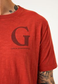 Camiseta de algodón roja con mangas cortas, cuello de canalé y logo negro que presenta una gran "G" y el texto "Garcia Jeanswear".