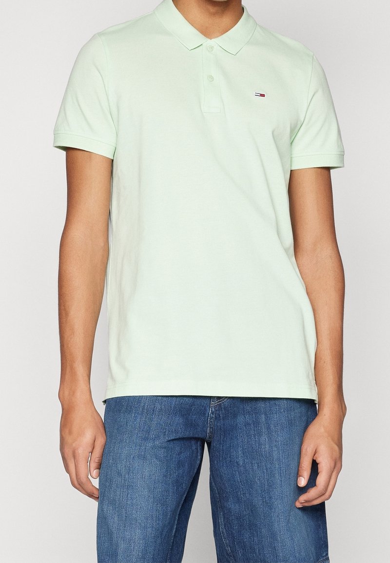Homme portant un polo à manches courtes vert clair et un jean bleu, debout les bras détendus le long du corps.
