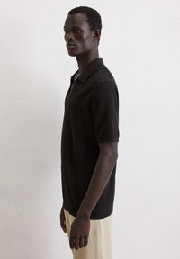 Polo noir �à manches courtes avec un tissu texturé, design à col ouvert et fentes sur les côtés, associé à un pantalon couleur crème.