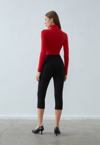 Rød langermet genser med høy hals, kombinert med svarte capri leggings og svarte høyhælte sko, sett fra baksiden mot en lys bakgrunn.