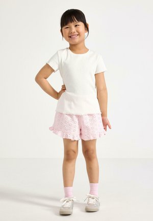 Hymyilevä tyttö, jolla on mustat hiukset, valkoinen lyhythihainen paita, vaaleanpunaiset pitsireunaiset shortsit, vaaleanpunaiset sukat ja harmaat lenkkarit, seisoo käsi lanteella.