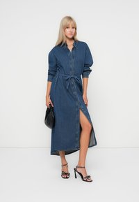 Denim jurk met lange mouwen, knopen aan de voorkant, strik in de taille, een kniehoge split en een rechte zoom. Draagt zwarte hakken en een zwarte tas.